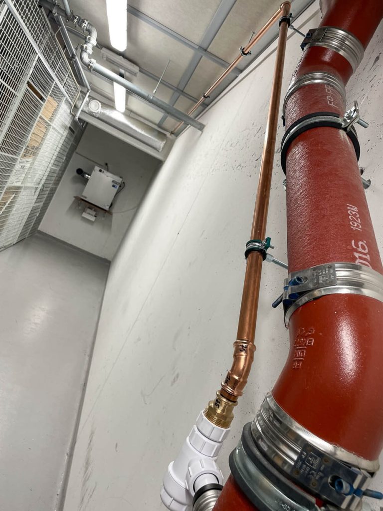 Et mørkerødt avløpsgrør i forgrunnen, med kobberrør parallelt som er koblet på nederst i bildekanten. En avfukter ses i bakgrunnen i korridoren i et bodareal.