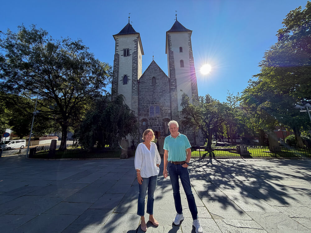 Hilde Brønlund og Odd Olav Fosso foran Mariakirken i Bergen, med sol og blå himmel i bakgrunnen.