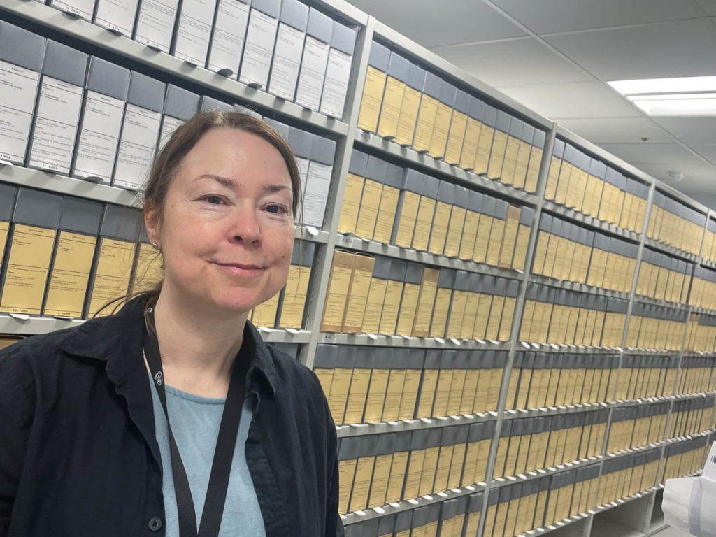 Mona Mårnes i arkivet hos Riksantikvaren, men rad på rad av dokumentbokser i bakgrunnen.