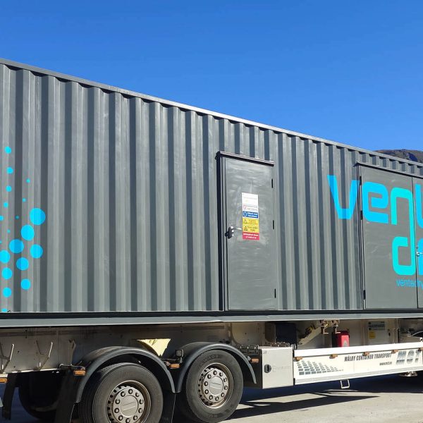 Mørk grå container med Ventadry-logo på lastebil med hvitt førerhus, sett mot blå himmel med fjell i brakgrunnen.
