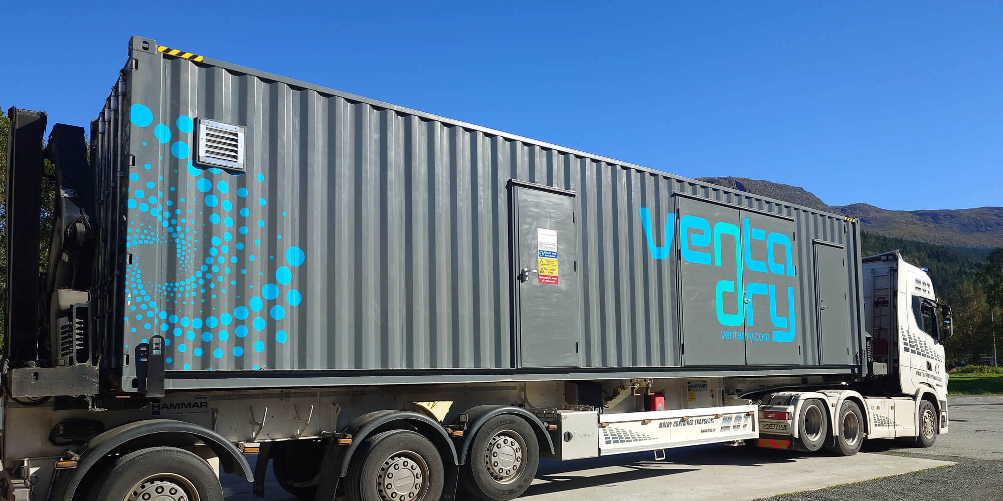 Mørk grå container med Ventadry-logo på lastebil med hvitt førerhus, sett mot blå himmel med fjell i brakgrunnen.