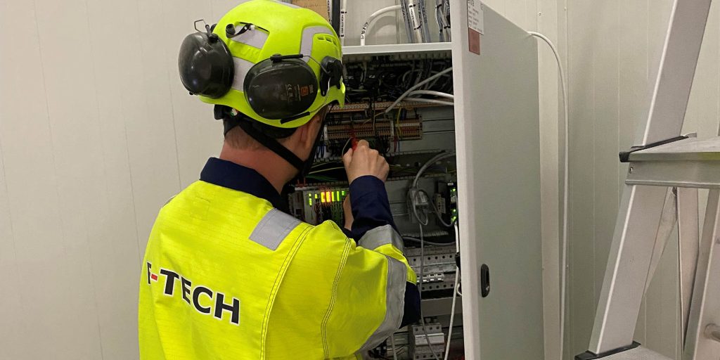 F-Techs automasjonsekspert med gult arbeidstøy og F-Tech logo, med ryggen til. jobber i koblingsskap.