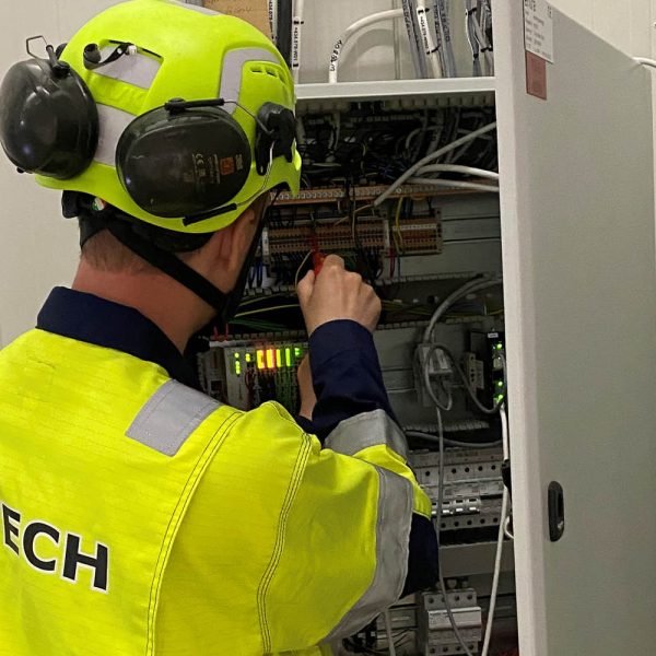 F-Techs automasjonsekspert med gult arbeidstøy og F-Tech logo, med ryggen til. jobber i koblingsskap.