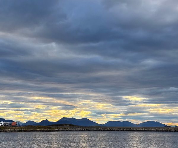 Hav , vei med lastebil mot blå fjell og delvis skyet himmel i solnedgang