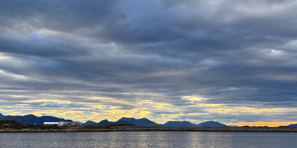 Hav , vei med lastebil mot blå fjell og delvis skyet himmel i solnedgang
